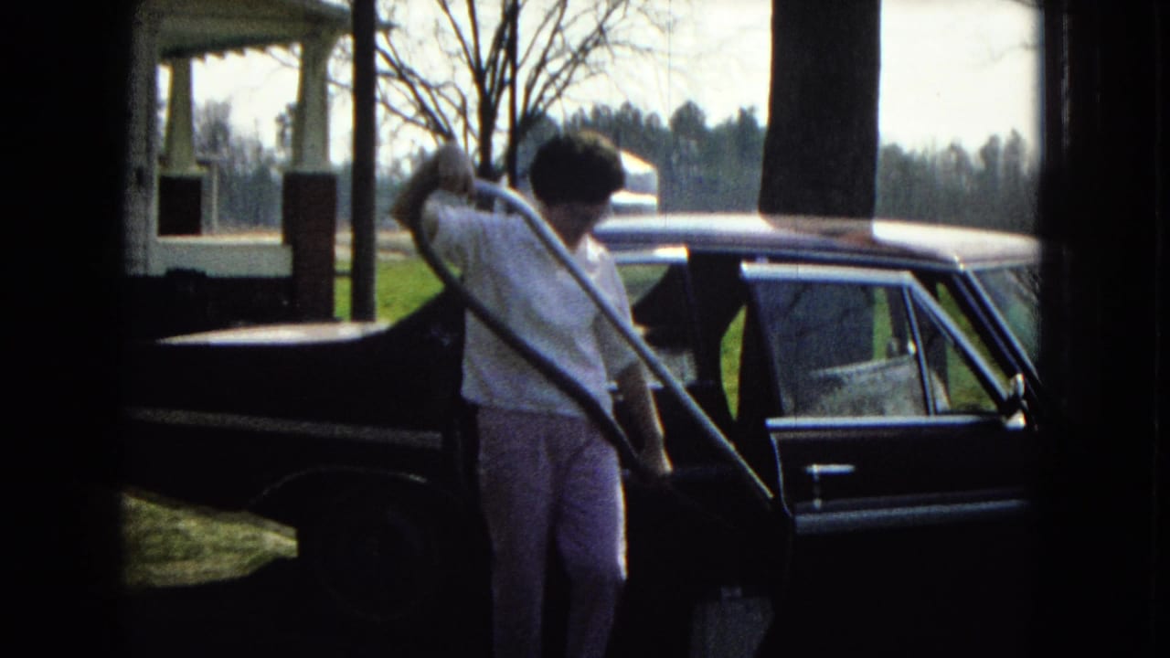 In a 1967 United States home-movie frame, a woman in a white shirt and light pants leans toward the open door of a dark car while using a vacuum hose near a house.