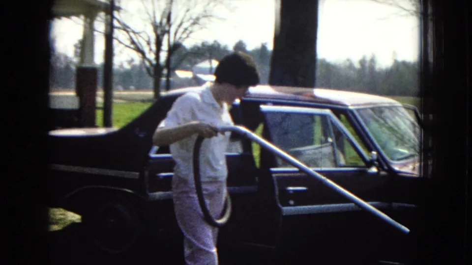 The woman stands beside the dark parked car with a vacuum hose looped in one hand and a long wand angled across the frame, with a porch and trees behind her.