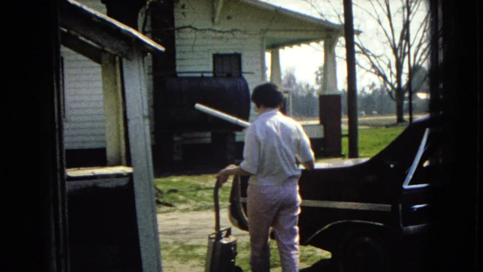 The woman moves along the driveway behind the dark car, carrying the vacuum hose near a white house with porch columns and a tank-like object beside it.