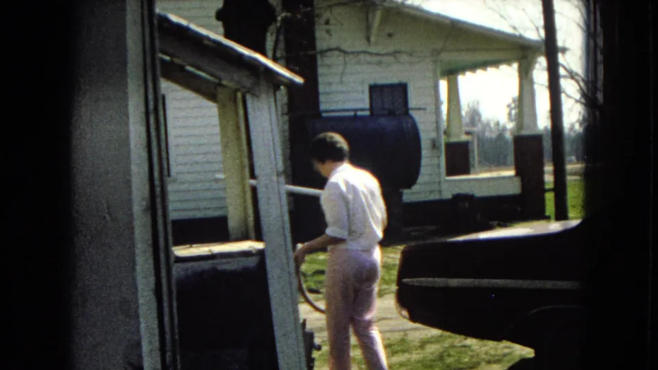 The woman continues vacuuming near the back of the dark car in a driveway, with the white house, porch, lawn, and bare trees visible in the background.