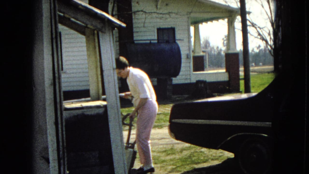 In 1967 United States 8mm footage, a woman in a white blouse and light pants bends with a vacuum hose near a dark sedan outside a white clapboard house.