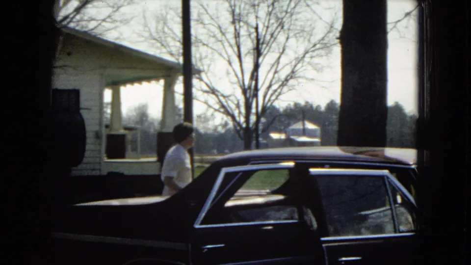 The woman moves along the side of the dark sedan near the white clapboard house, with the car roofline, chrome trim, lawn, and trees filling the frame.