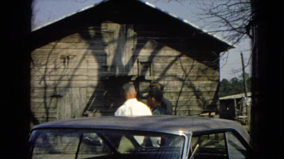 Two adult men stand near a weathered wooden building behind the roof of a parked car, expanding the scene from car cleaning to nearby residential activity.