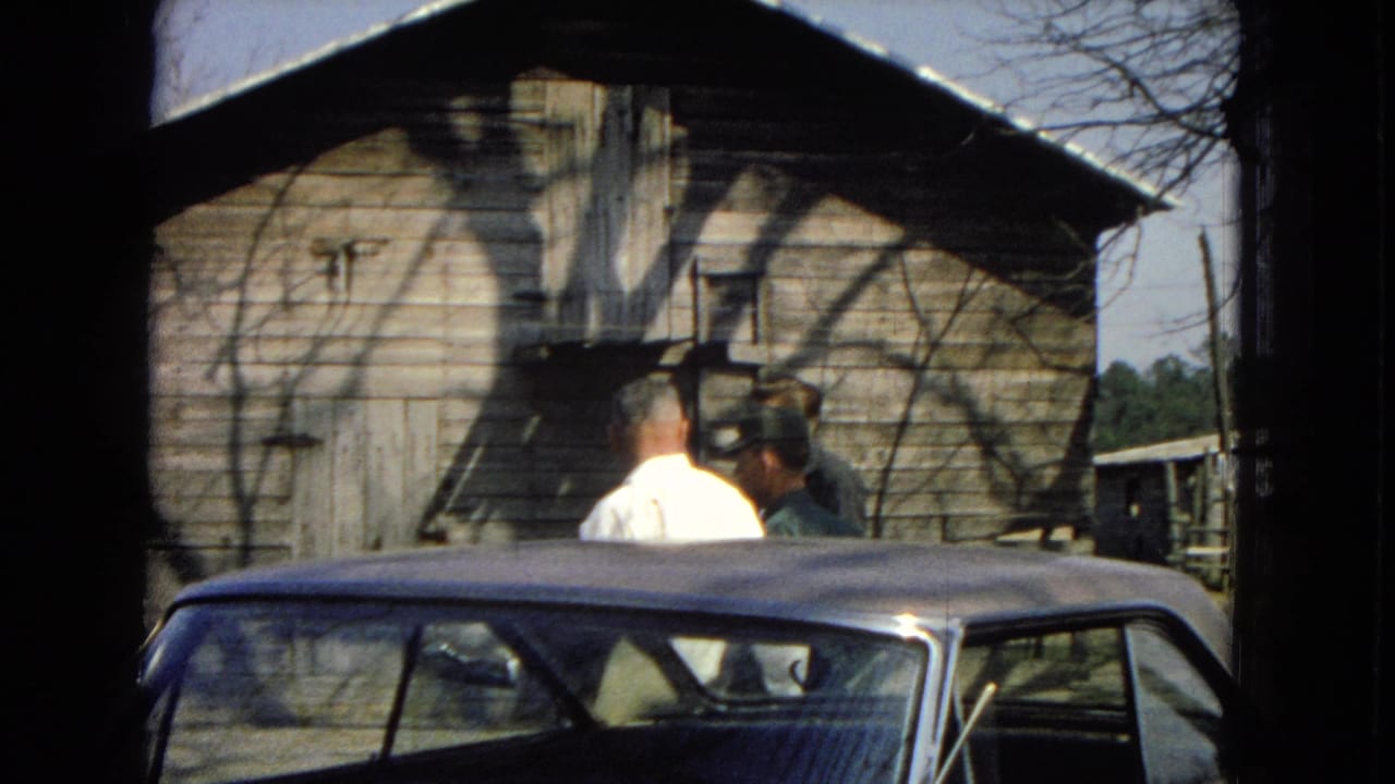 Three adult men converse beside a classic car in a rural United States home-movie scene from 1967, with a weathered wooden barn behind them.