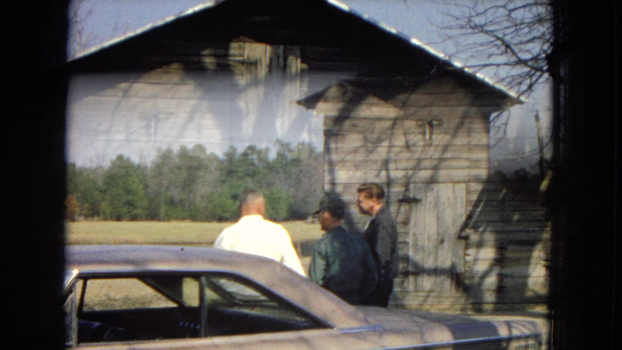 Three men talk next to a classic sedan with a barn and field behind them.