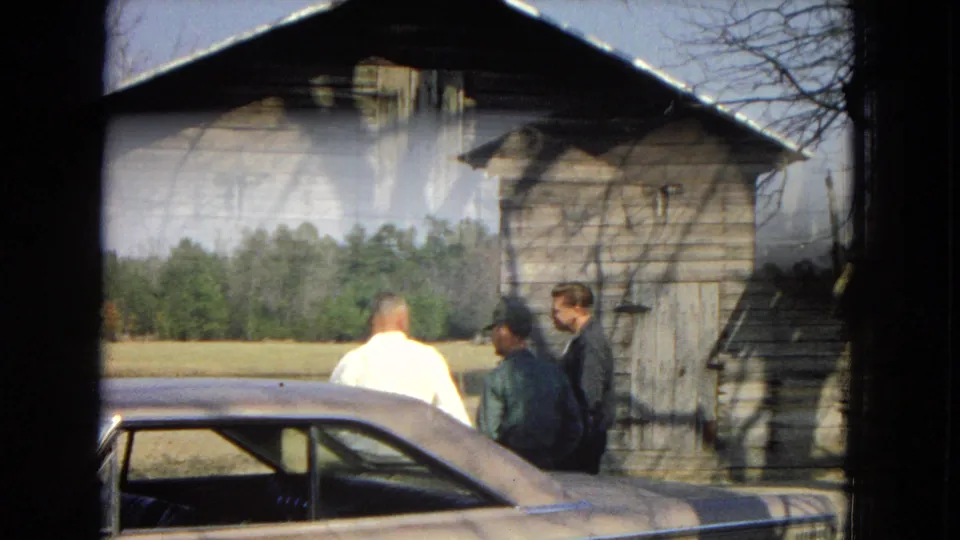Three adult men stand by a classic sedan in a rural 1967 United States home movie, with a barn, field, and tree line visible beyond the car.