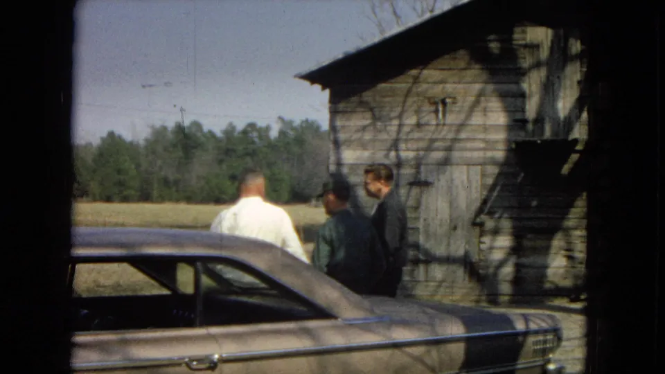 Three men remain in conversation beside a classic car, with the barn wall, open field, and distant trees visible in the 1967 rural home-movie frame.