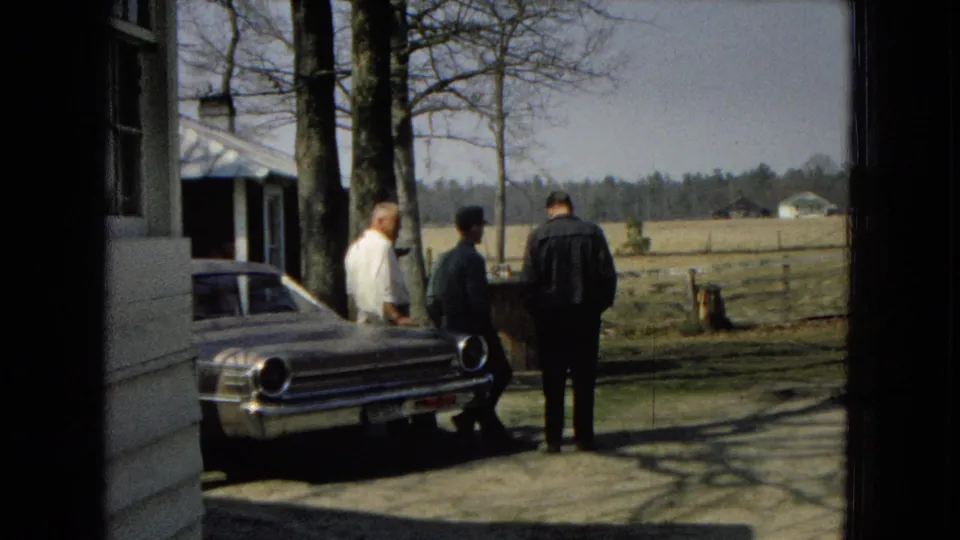 Three adult men are seen near the rear of a classic car by a farmhouse and trees, with fenced fields stretching into the background in the 1967 home-movie footage.