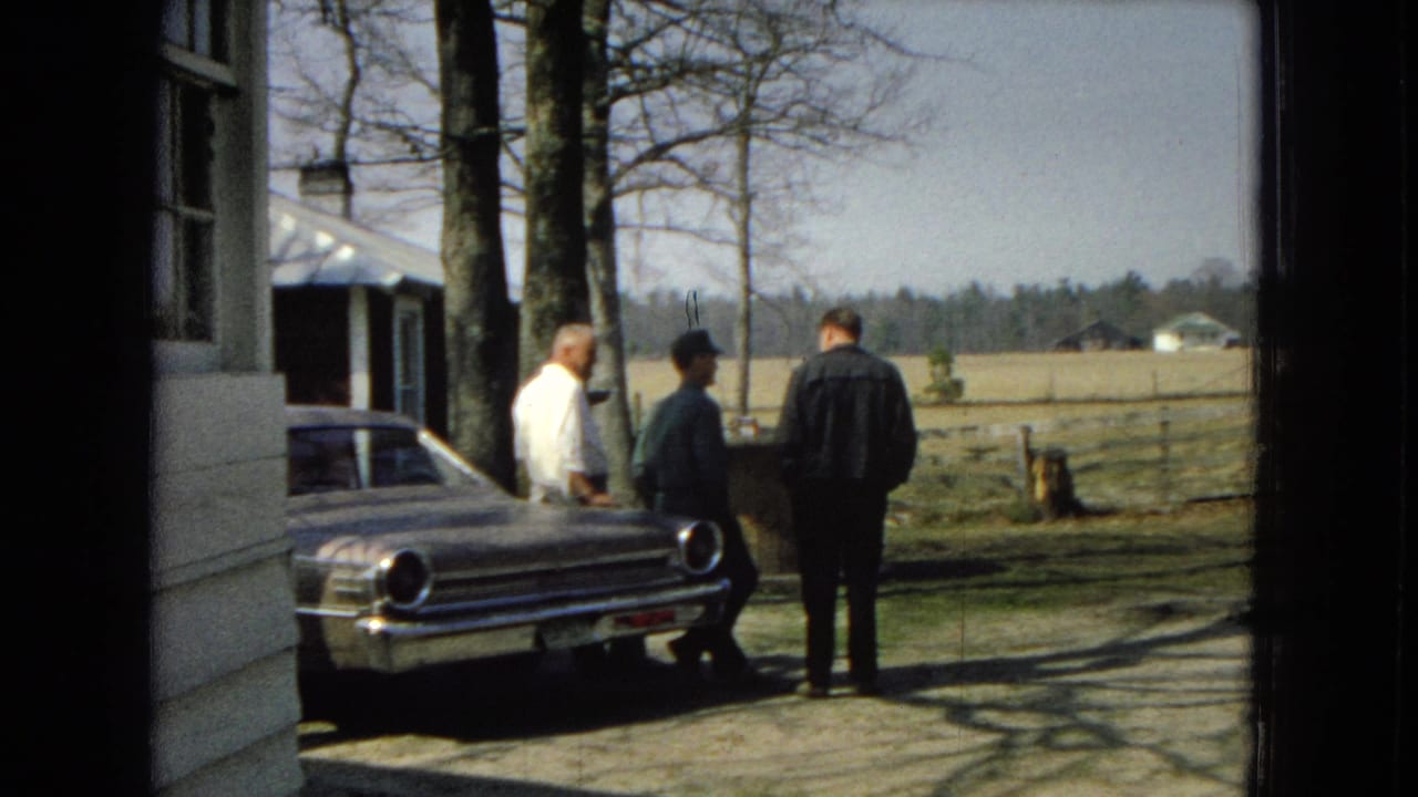 Three adult men remain beside the sedan near a farmhouse, with bare trees, fencing, and fields visible beyond them.