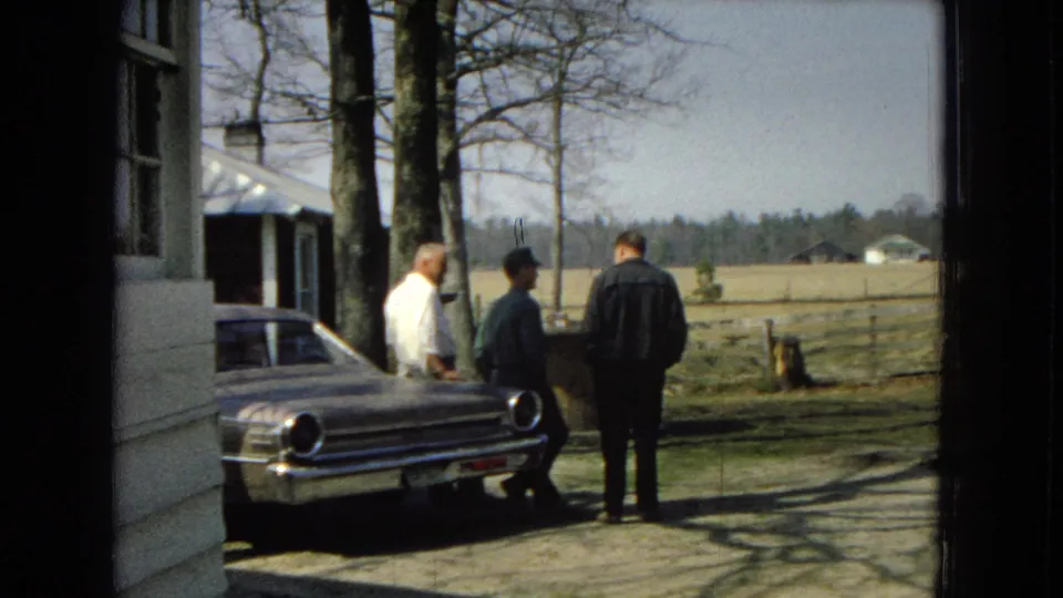 Three adult men remain beside the sedan near a farmhouse, with bare trees, fencing, and fields visible beyond them.