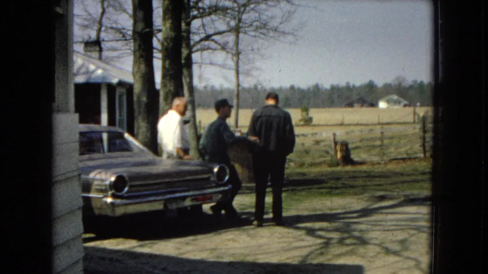 Three adult men linger by a parked chrome sedan outside a farmhouse, with rural fencing and distant buildings visible in the background.