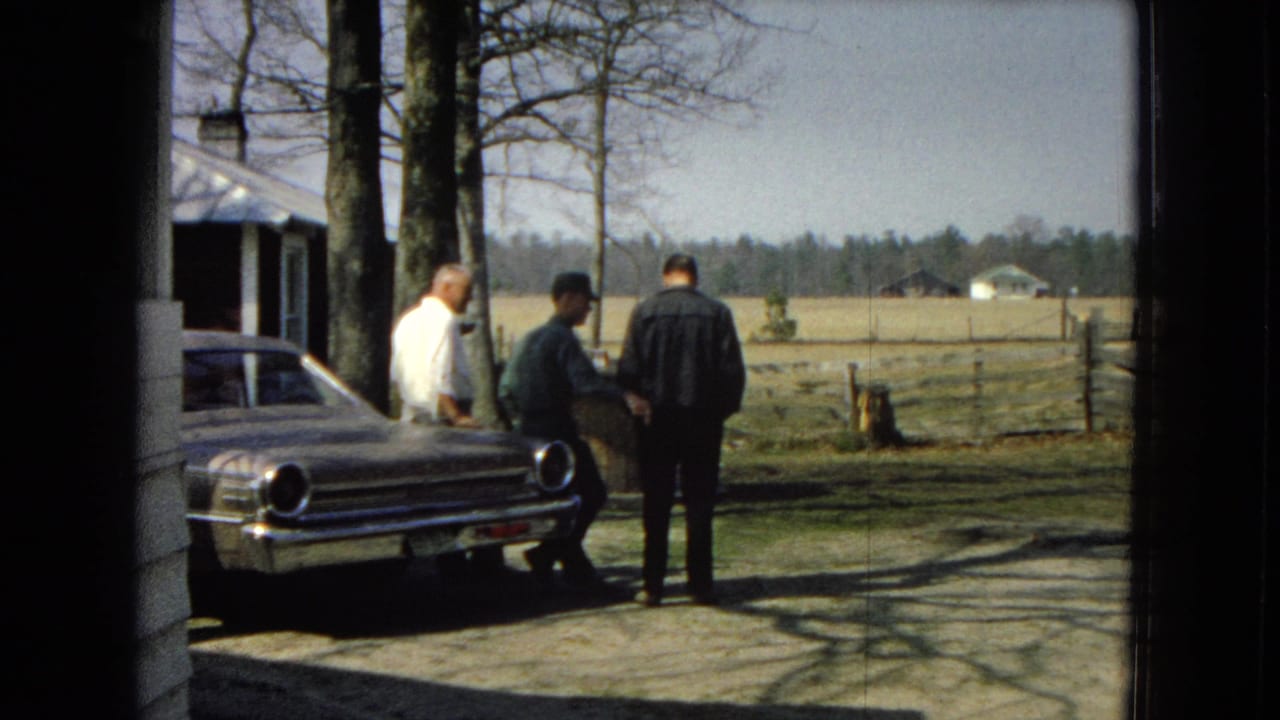 Three men stand beside a light-colored sedan in a rural driveway.