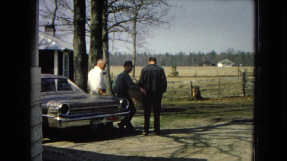 Three adult men stand near a light-colored mid-century sedan in a rural United States driveway, with trees, fencing, open fields, and a building edge visible.
