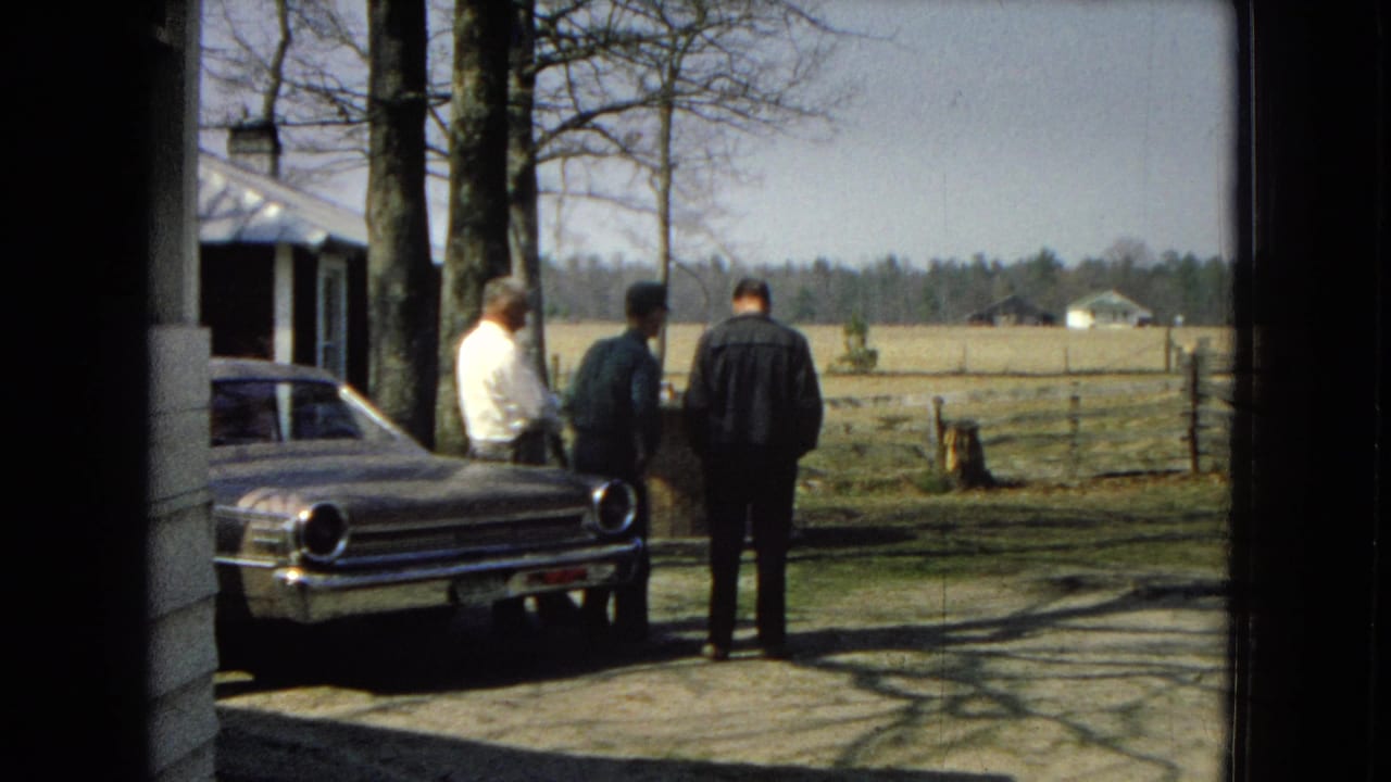 Three men face a parked sedan near trees and open fields.