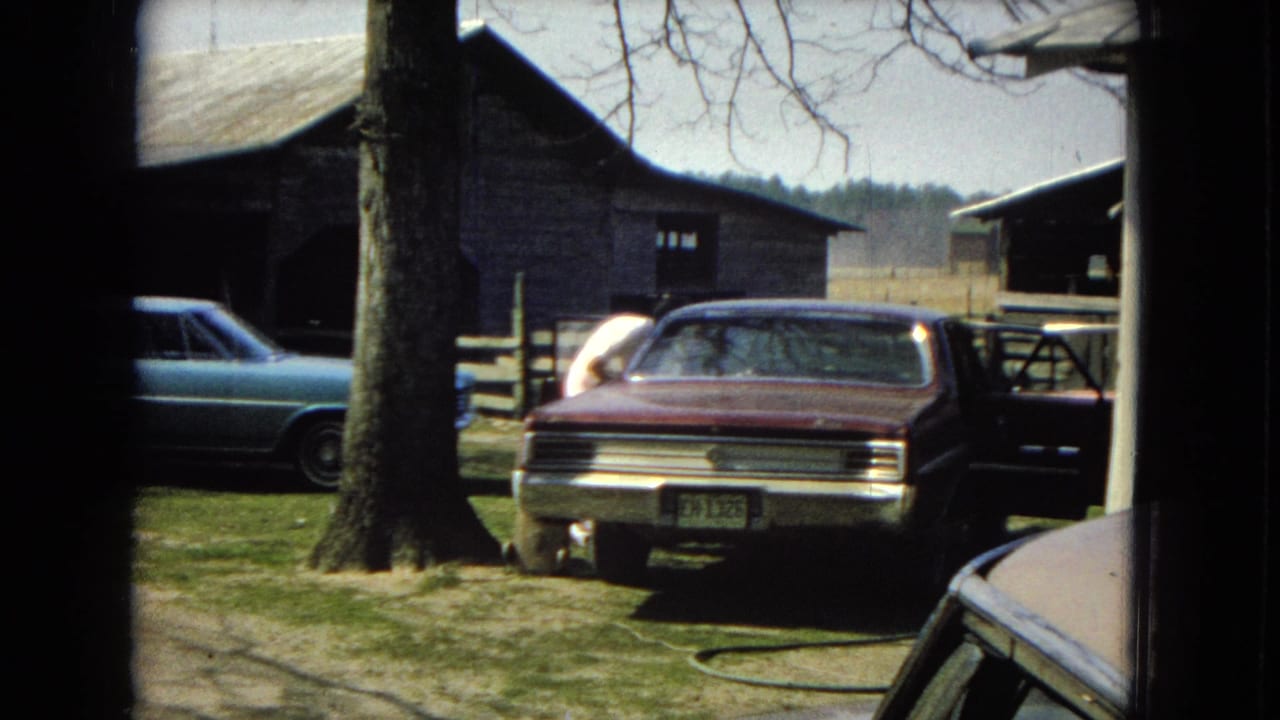 A rural United States yard scene from the supplied 1967 home-movie metadata shows a maroon sedan near a large tree, a blue car at left, and weathered wooden barn structures in the background.