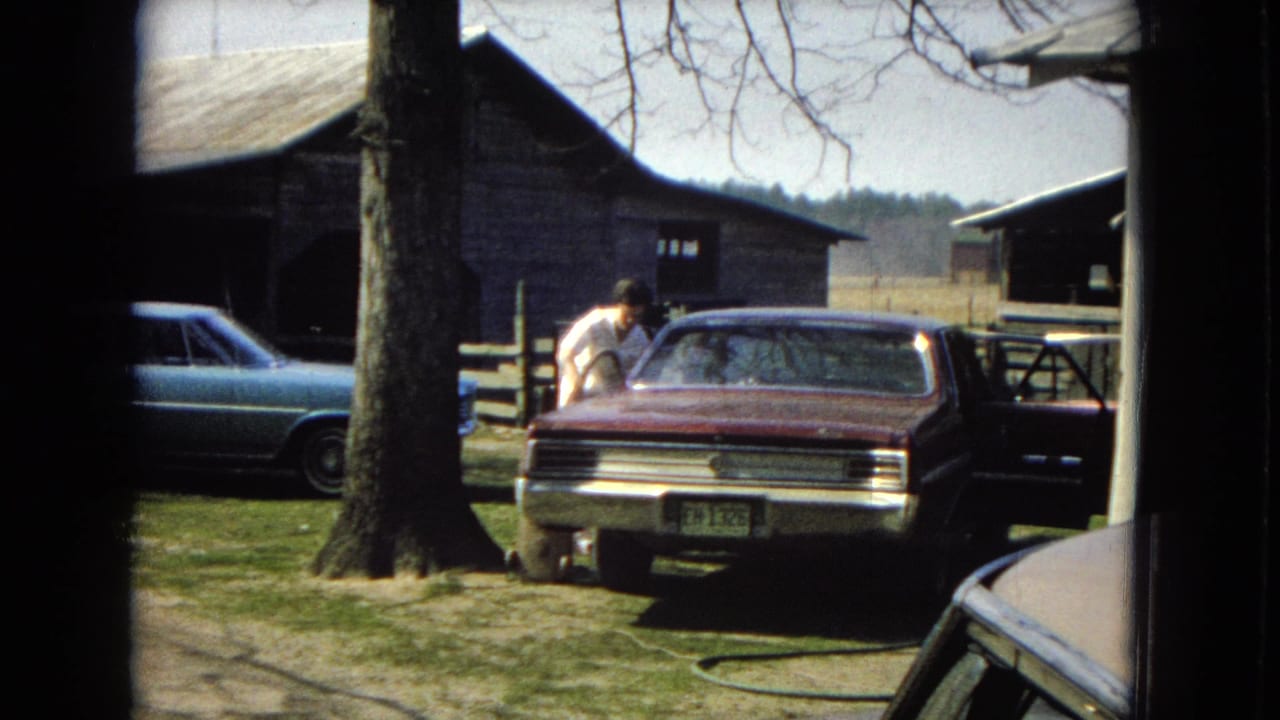Man in a light shirt leans near the rear side of a maroon sedan in a rural yard.