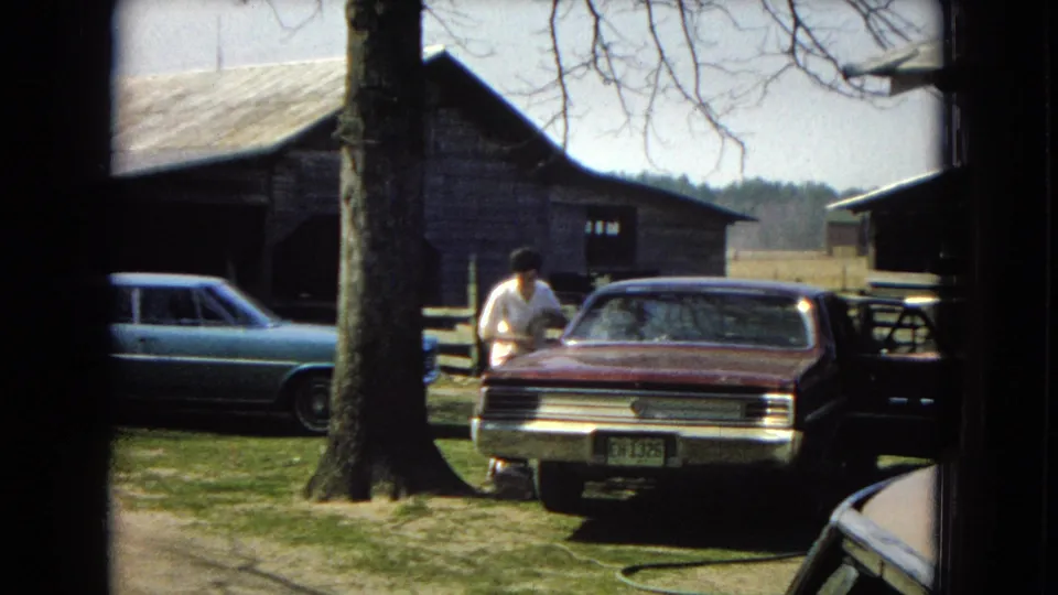 At the end of the clip, the man stands beside the maroon sedan in the sunny yard, with a hose or cord on the grass, a blue car at left, and barn structures behind him.