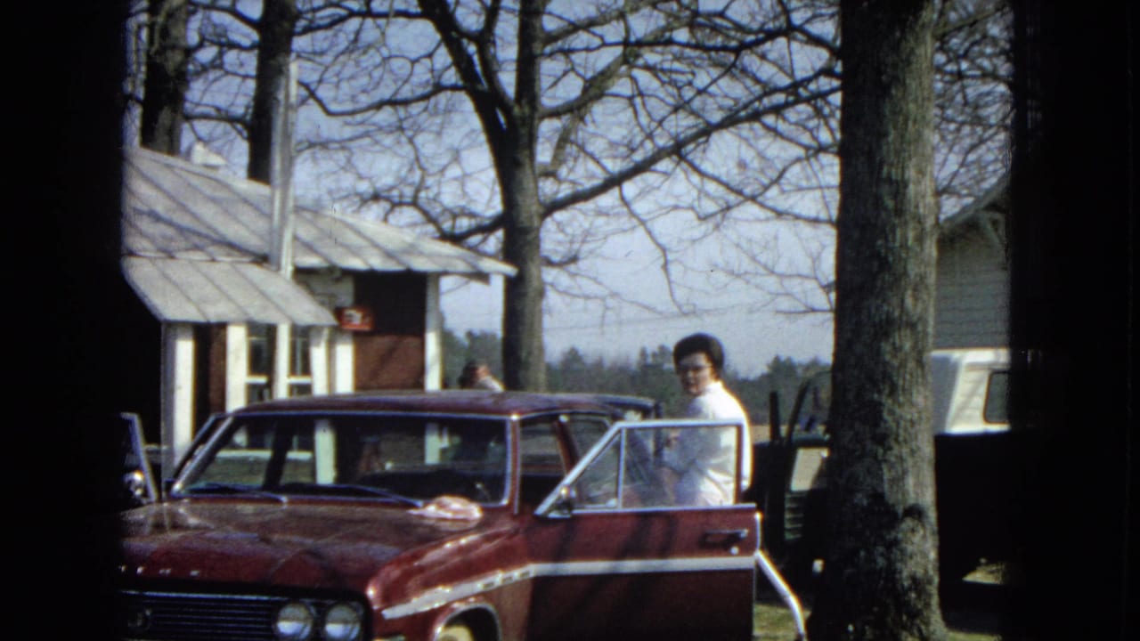 In a 1967 United States home-movie frame, a woman in a white shirt stands by the open door of a maroon sedan near bare trees and modest residential houses.