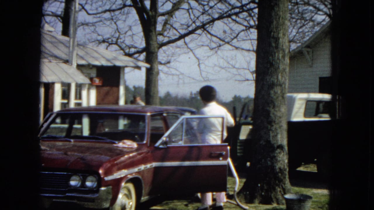 Woman leaning near the open door of a maroon sedan with houses and a white truck behind her.