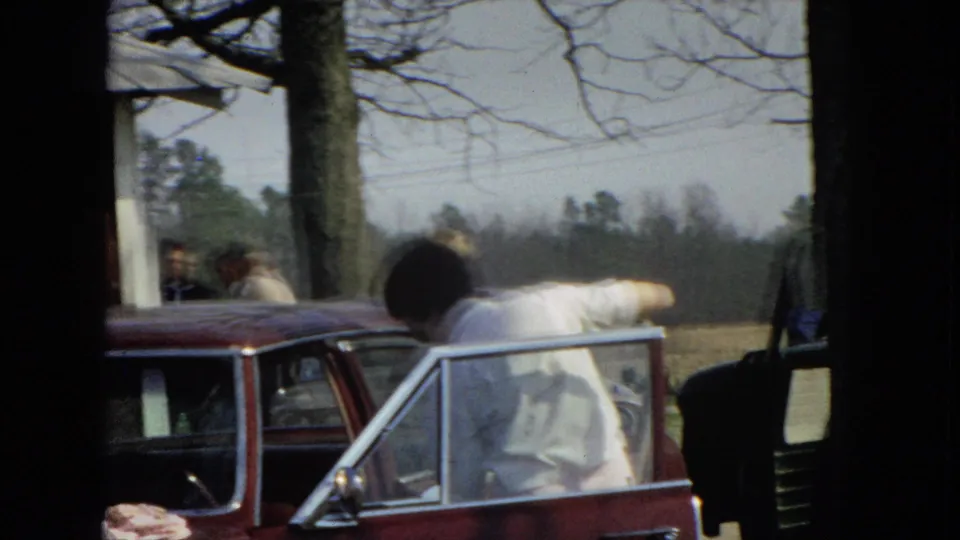 The woman leans into or across the open door area of the maroon sedan, with trees, grass, and another person partially visible in the residential yard.