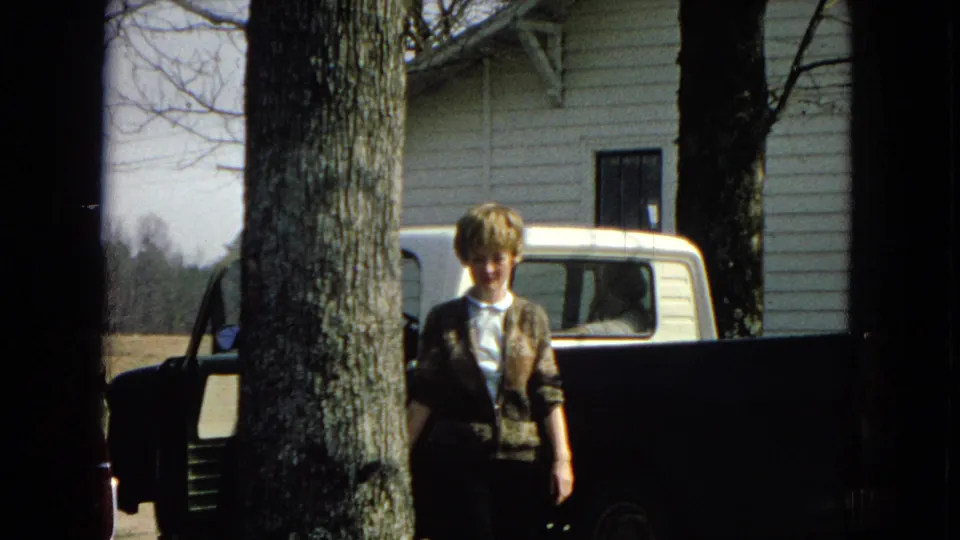 A blond person stands near a tree in front of a white pickup truck and house exterior, while the maroon car remains partly visible at the edge of the frame.