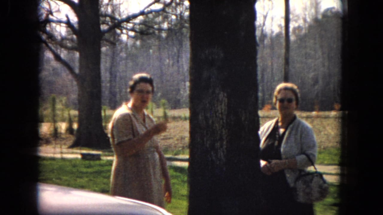 In 1967 United States 8mm footage, two adult women stand in a sunny wooded or landscaped area as one gestures while speaking and a car hood appears at the lower edge.