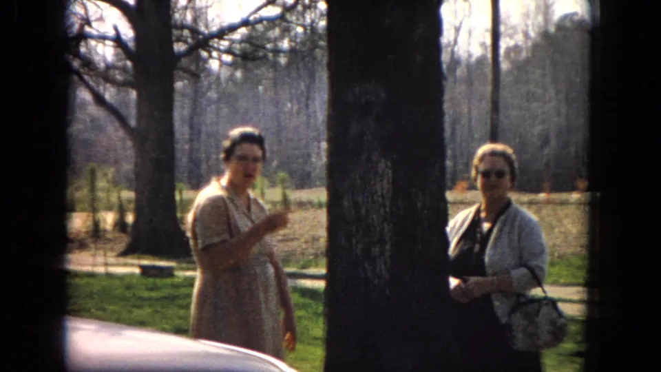 In 1967 United States 8mm footage, two adult women stand in a sunny wooded or landscaped area as one gestures while speaking and a car hood appears at the lower edge.