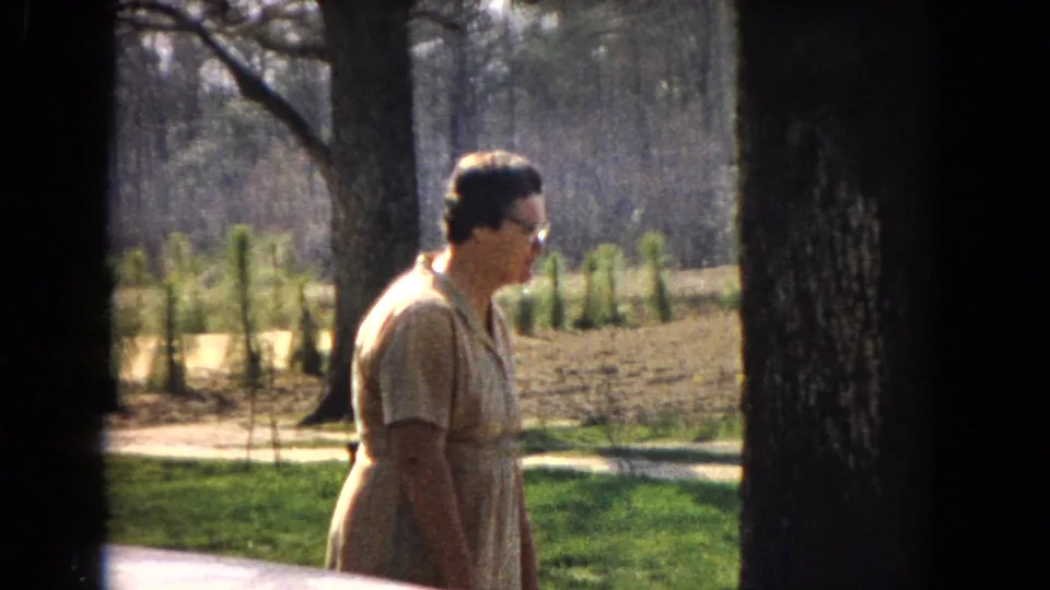A woman in a light-colored dress and glasses appears in profile in 1967 United States 8mm footage, standing or moving near tree trunks, grass, and a paved path.