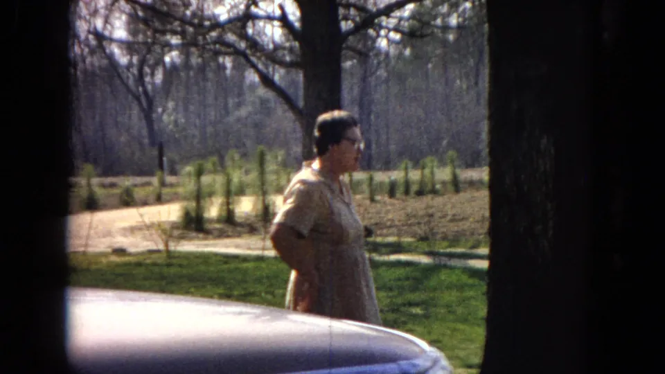 An adult woman in a light dress stands in profile near the edge of a car hood in 1967 United States home-movie footage, with tree trunks and landscaped grass in the background.