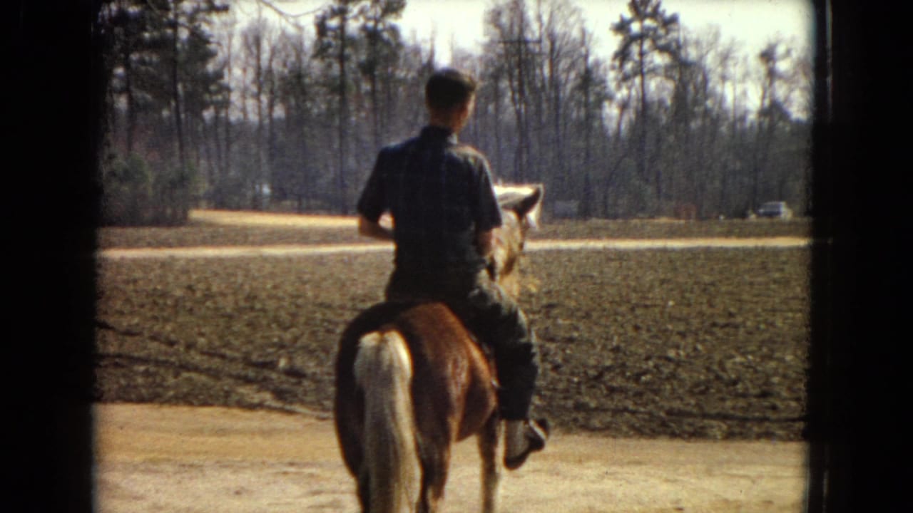 In 1967 United States home-movie footage, a boy rides a brown pony with a pale tail along a dirt path beside a plowed field and leafless trees.