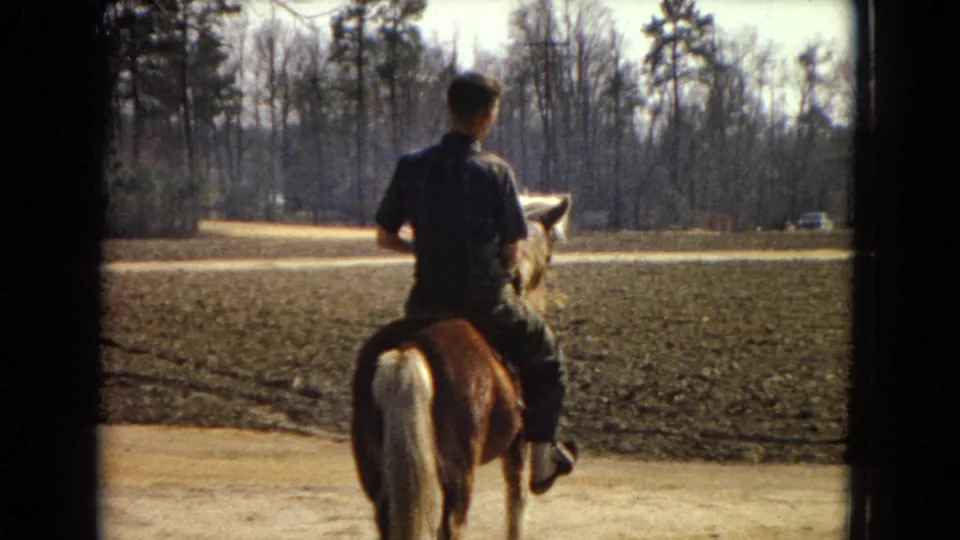 In 1967 United States home-movie footage, a boy rides a brown pony with a pale tail along a dirt path beside a plowed field and leafless trees.