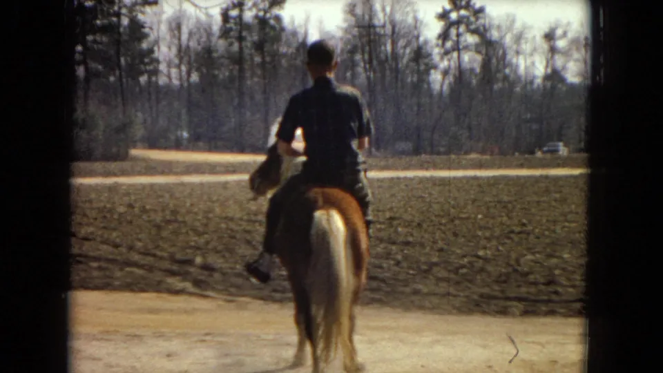 The rider continues away from the camera on a brown pony, with open plowed ground, a pale dirt road, bare trees, and a small distant car visible beyond the field.