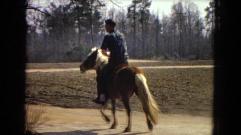 A side view shows the boy riding the pony at a trot along the dirt path, with the plowed field and leafless tree line behind him in the 8mm frame.