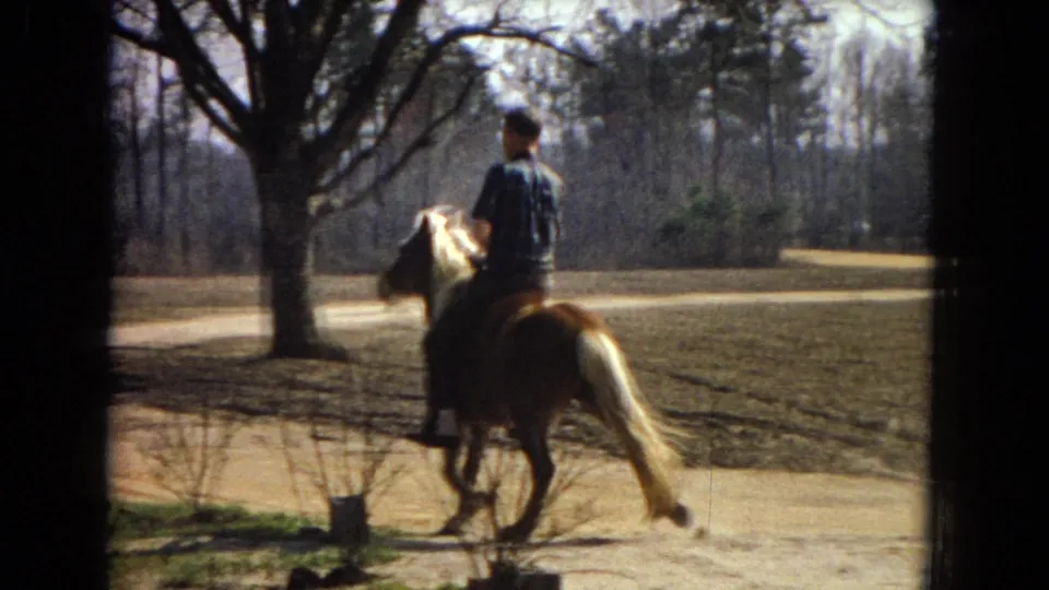 The pony and rider move past a large tree near the field edge, with a dirt path, plowed ground, bare trees, and soft 8mm color texture visible.