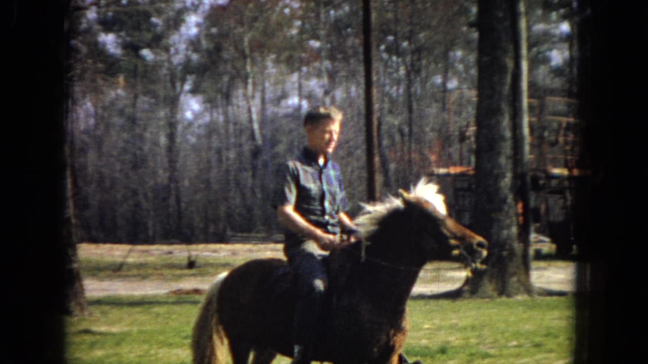 In 1967 Arizona home-movie footage, a young man in a short-sleeved plaid shirt rides a saddled brown horse with a white mane through a grassy, tree-lined property.