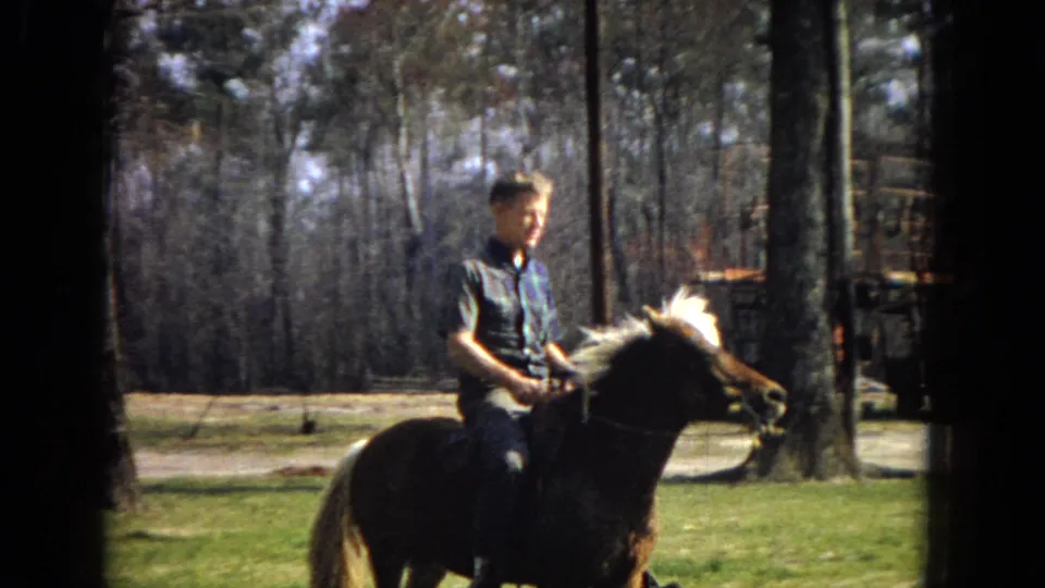 In 1967 Arizona home-movie footage, a young man in a short-sleeved plaid shirt rides a saddled brown horse with a white mane through a grassy, tree-lined property.