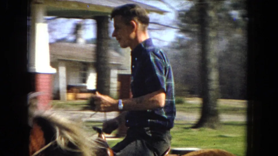 A side view shows the young man in a blue plaid short-sleeved shirt holding the reins while riding past a porch area, trees, and grass in the 1967 home-movie scene.