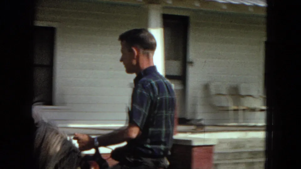 The man rides beside a white house or porch structure, seen in profile as the horse moves close to the building in this 1967 Arizona 8mm home-movie frame.