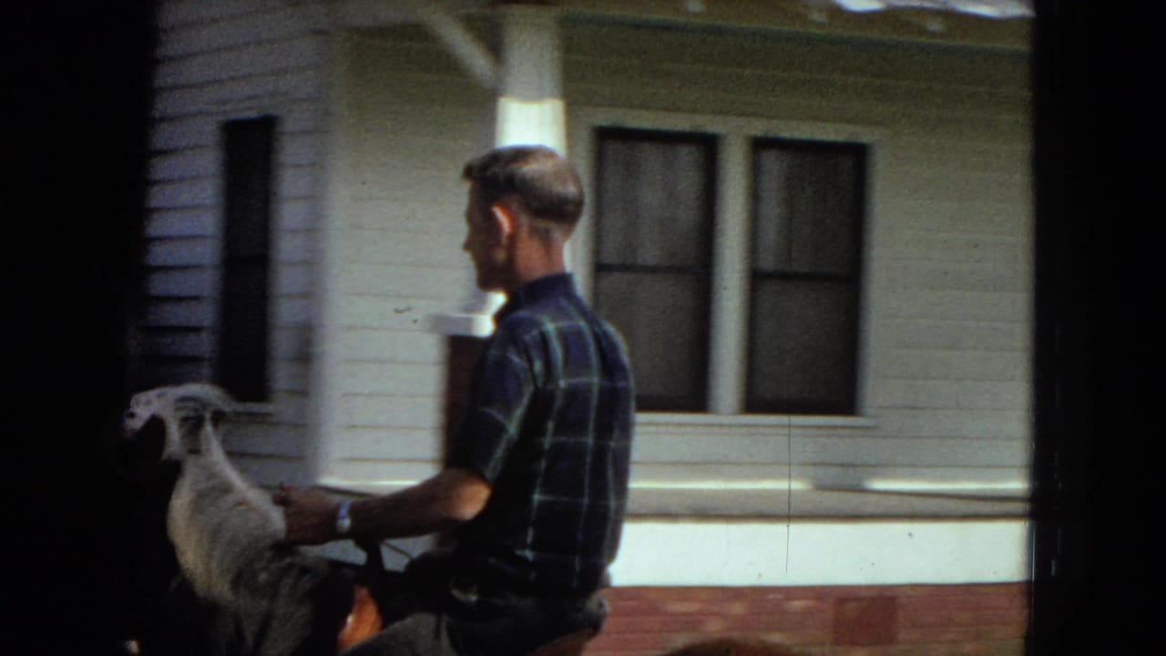1967 home-movie frame showing an adult man in a blue plaid shirt riding a light-colored horse beside a white-sided house with porch columns.