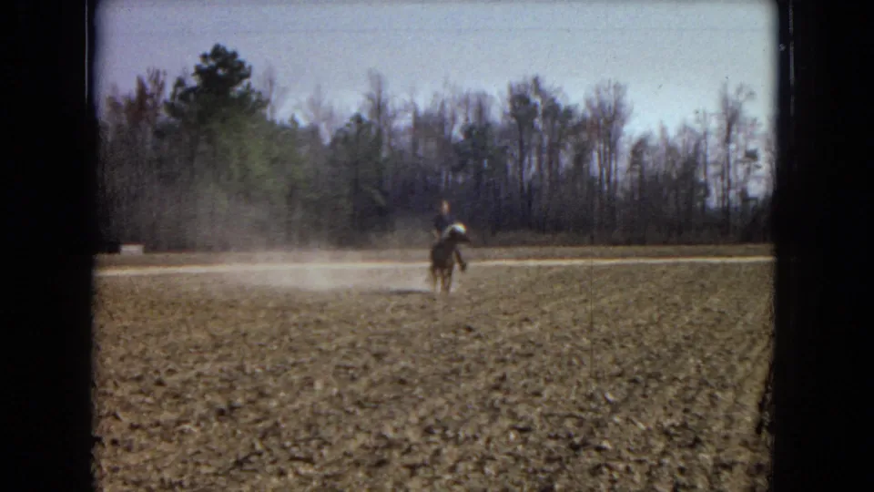 1967 home-movie frame showing a rider on horseback in a wide plowed field, with a low dust cloud and bare trees beyond.