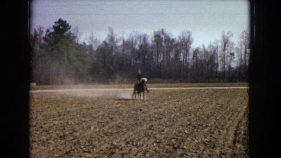 1967 home-movie frame showing the rider and horse small in the frame as they continue across the rural field with a wooded edge behind them.