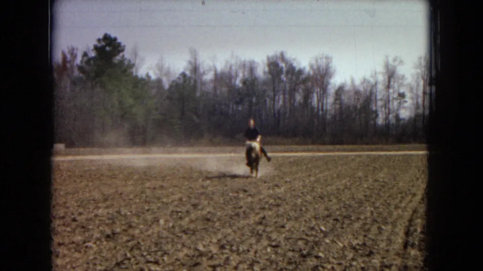The rider guides a small pony toward the camera across rough plowed ground, with a tree line and dust plume visible in the rural Arizona setting.