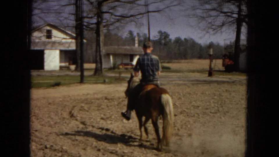 A rider in a plaid shirt is seen from behind on a small pony near a pale outbuilding, with trees, dirt ground, and open rural space around them.