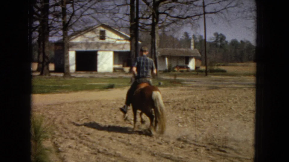 The rider continues away on the pony beside a pale rural building with an open doorway, bare trees, dusty ground, and a shadow trailing behind.