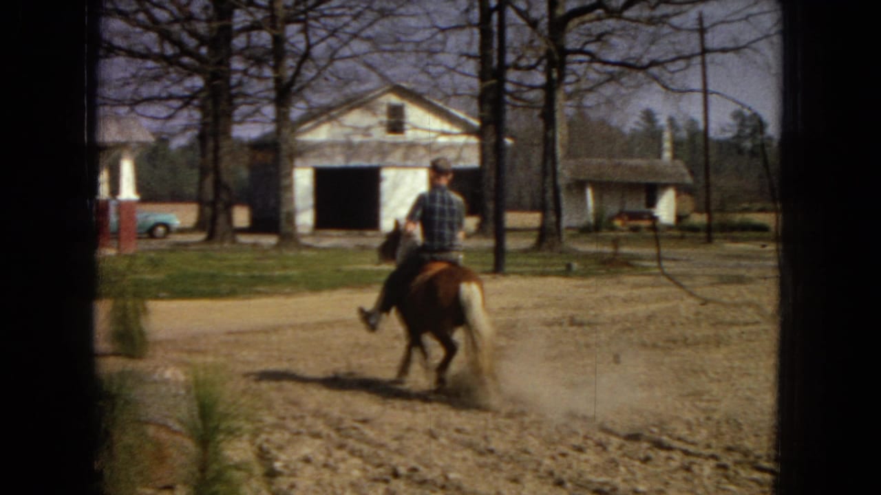 In supplied 1967 Arizona home-movie footage, a man rides a small brown horse across a dusty yard with a white farmhouse, detached garage, bare trees, and a light blue car in the background.