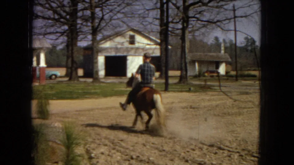 In supplied 1967 Arizona home-movie footage, a man rides a small brown horse across a dusty yard with a white farmhouse, detached garage, bare trees, and a light blue car in the background.