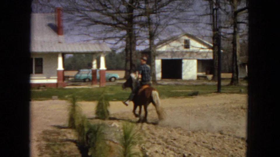 The rider guides the small brown horse past a white farmhouse with red brick porch columns, a detached garage, bare trees, and parked light blue cars visible in the yard area.