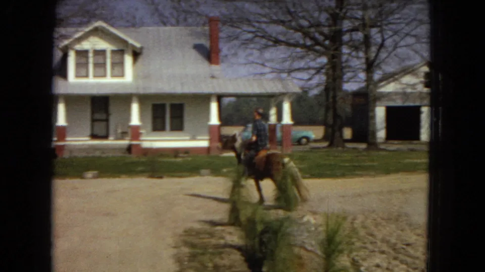 The home-movie frame shows the horse and rider partly aligned with young shrubs in the foreground, with the white house, porch columns, garage, car, and bare trees behind them.