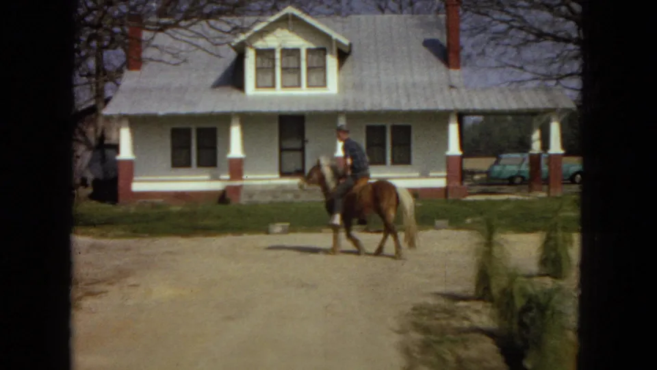 At the end of the short clip, the man rides the small horse across the open yard in front of the white farmhouse, with a light blue car, bare trees, low shrubs, and dirt roadway visible.