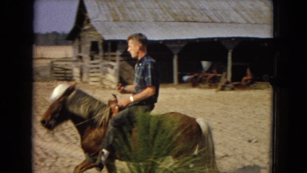 In 1967 United States home-movie footage, an adult man in a plaid short-sleeve shirt rides a small brown pony with a pale mane past a weathered barn and wooden fencing.